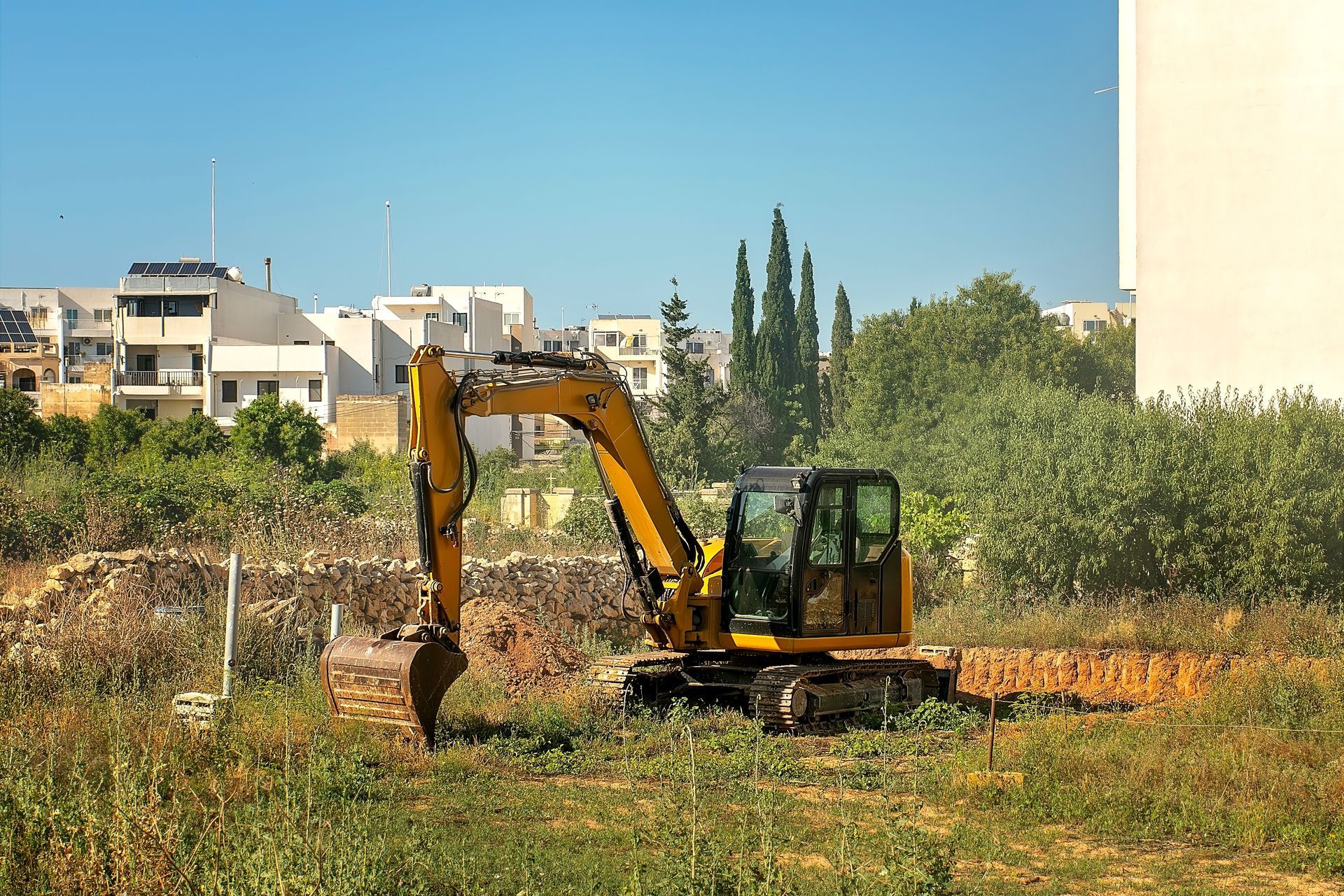 A yellow excavator sits in a grassy, undeveloped lot with residential buildings in the background.