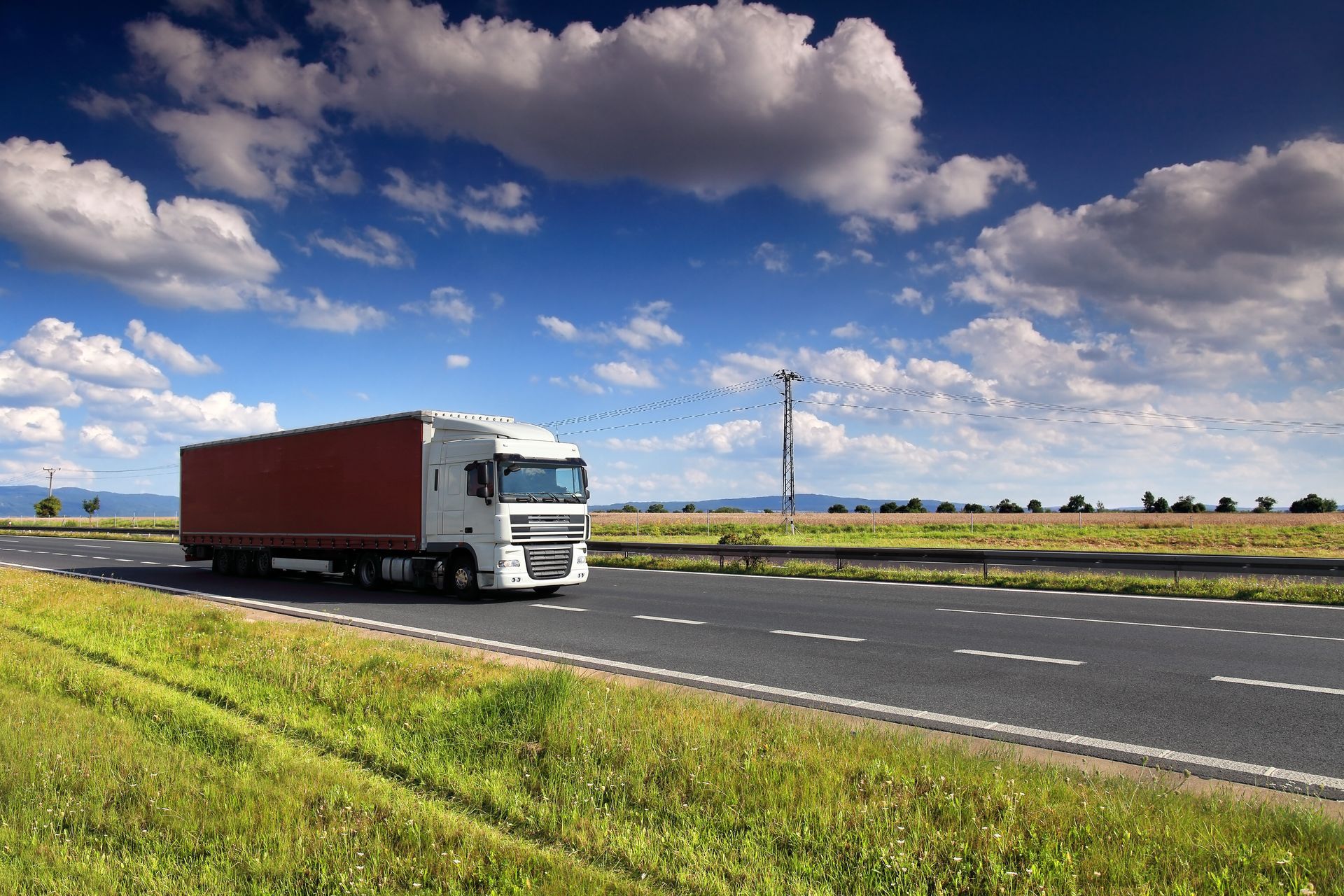 A white semi-truck with a long, red trailer driving along an open highway under a blue, cloudy sky.