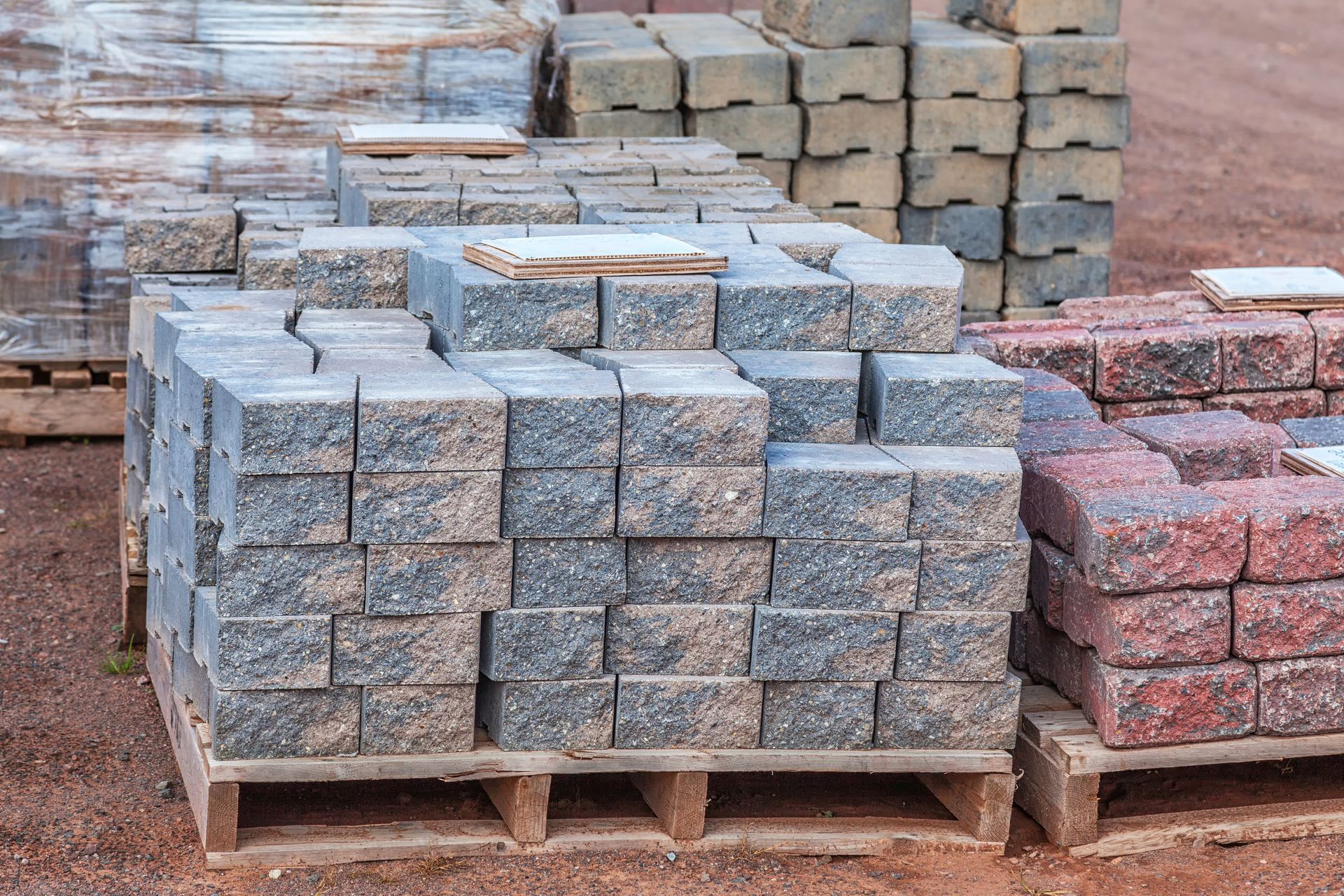 Wooden pallets stacked with grey and reddish-brown rectangular concrete pavers on a dirt surface.