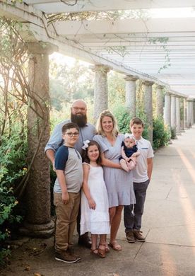 Family of six poses under a stone pergola. Father with beard, mother in blue dress, children smiling.