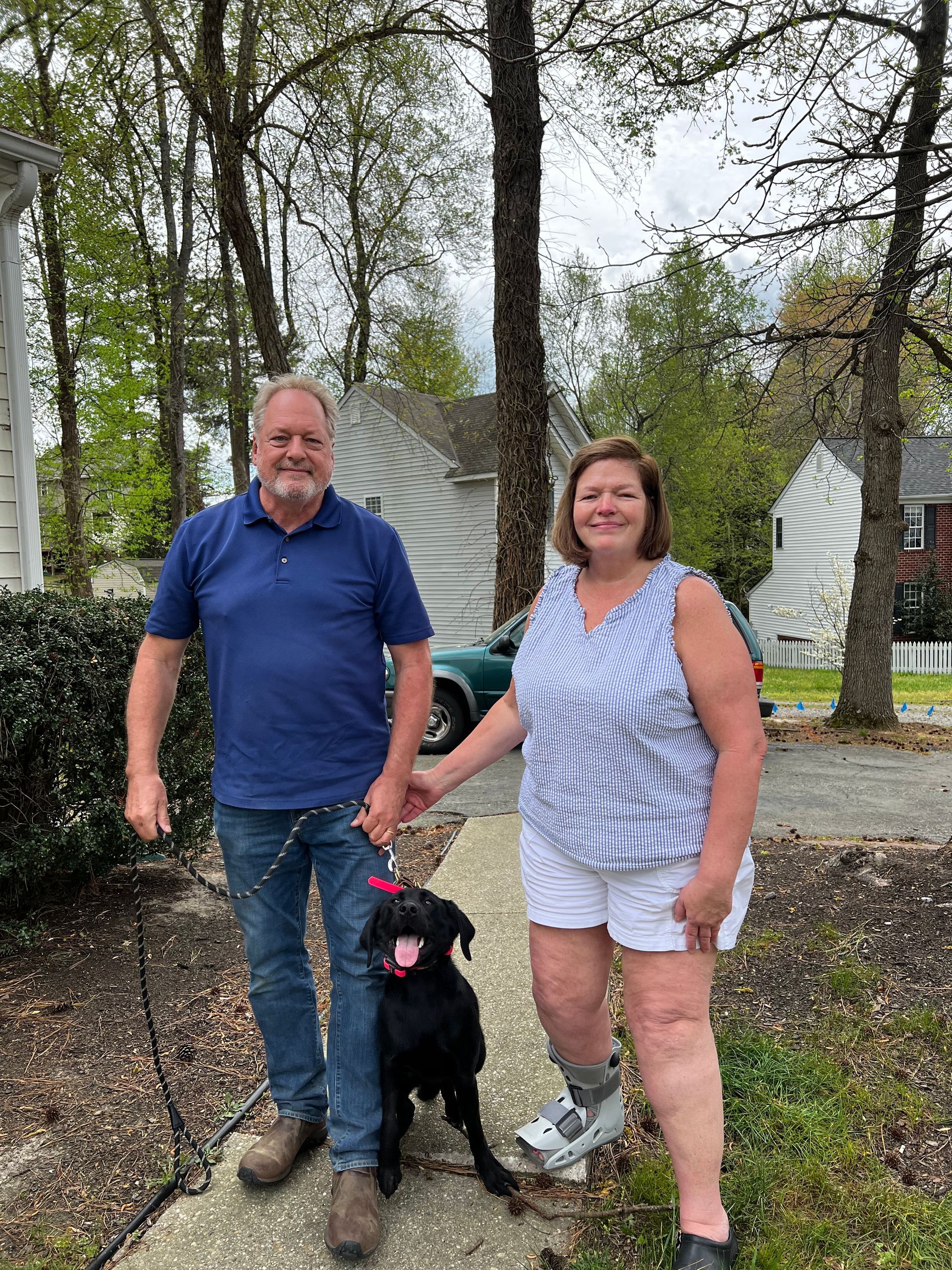 Man and woman holding hands, walking black lab on a leash in front of a house.