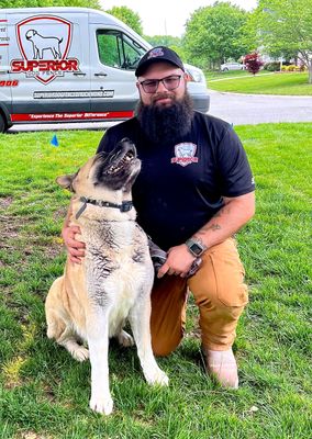 A man with a beard is kneeling down next to a dog in the grass.