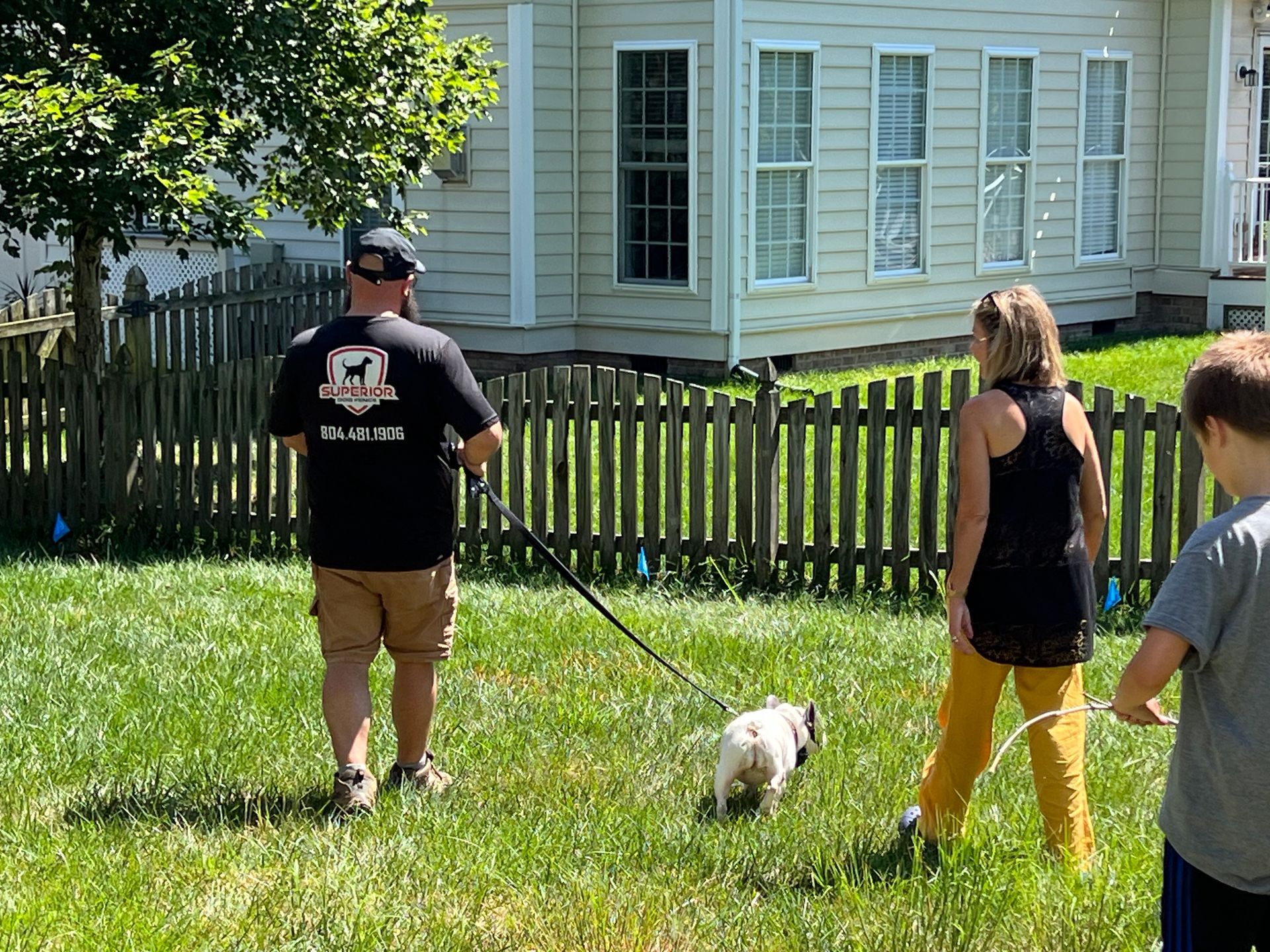 Man walking a pug on a leash with a woman and child in a yard near a house and fence.