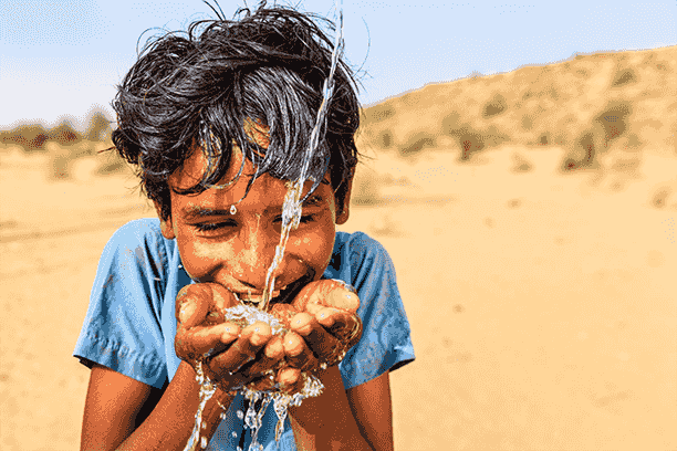 A young boy is drinking water from his hands in the desert.