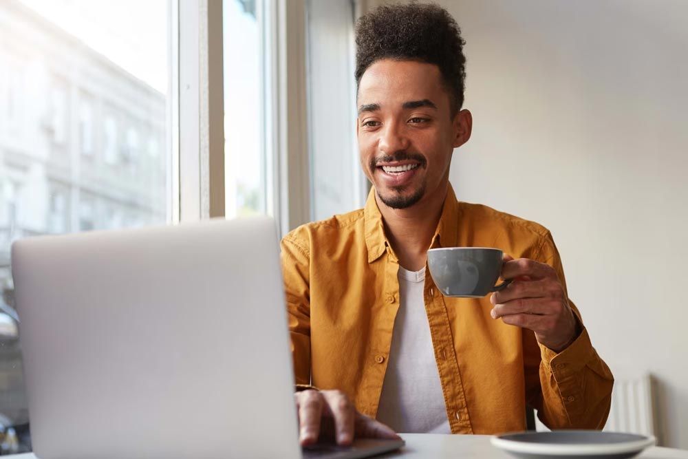 A man is sitting at a table using a laptop computer while holding a cup of coffee.