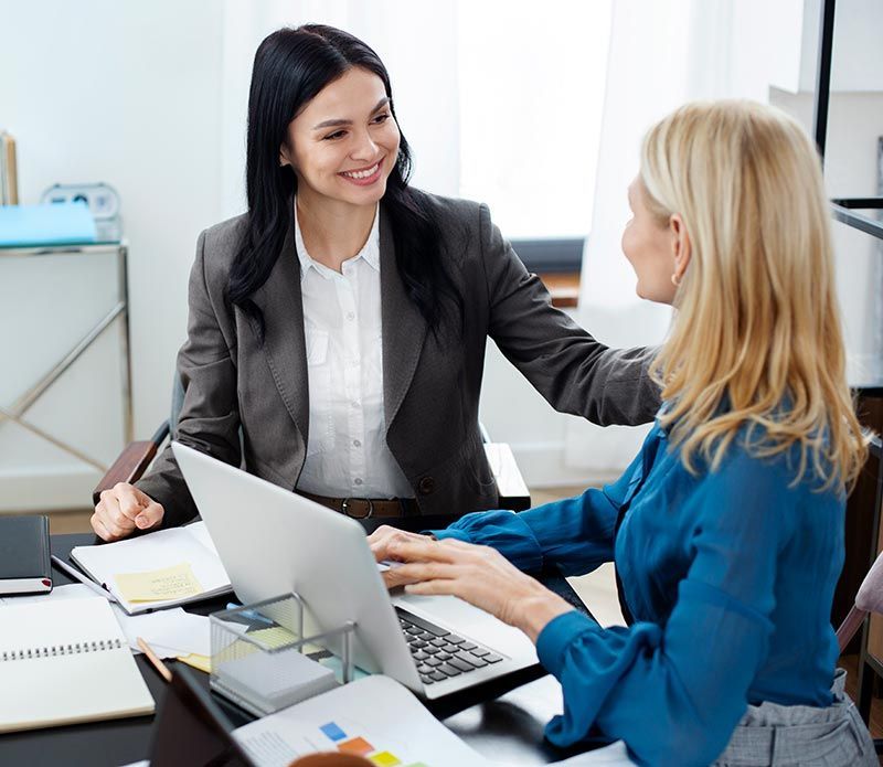Two women are sitting at a desk with a laptop and talking to each other.