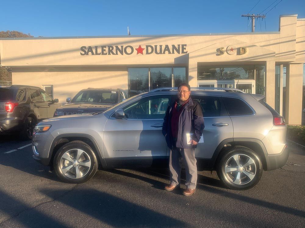A man is standing in front of a silver jeep at a car dealership.
