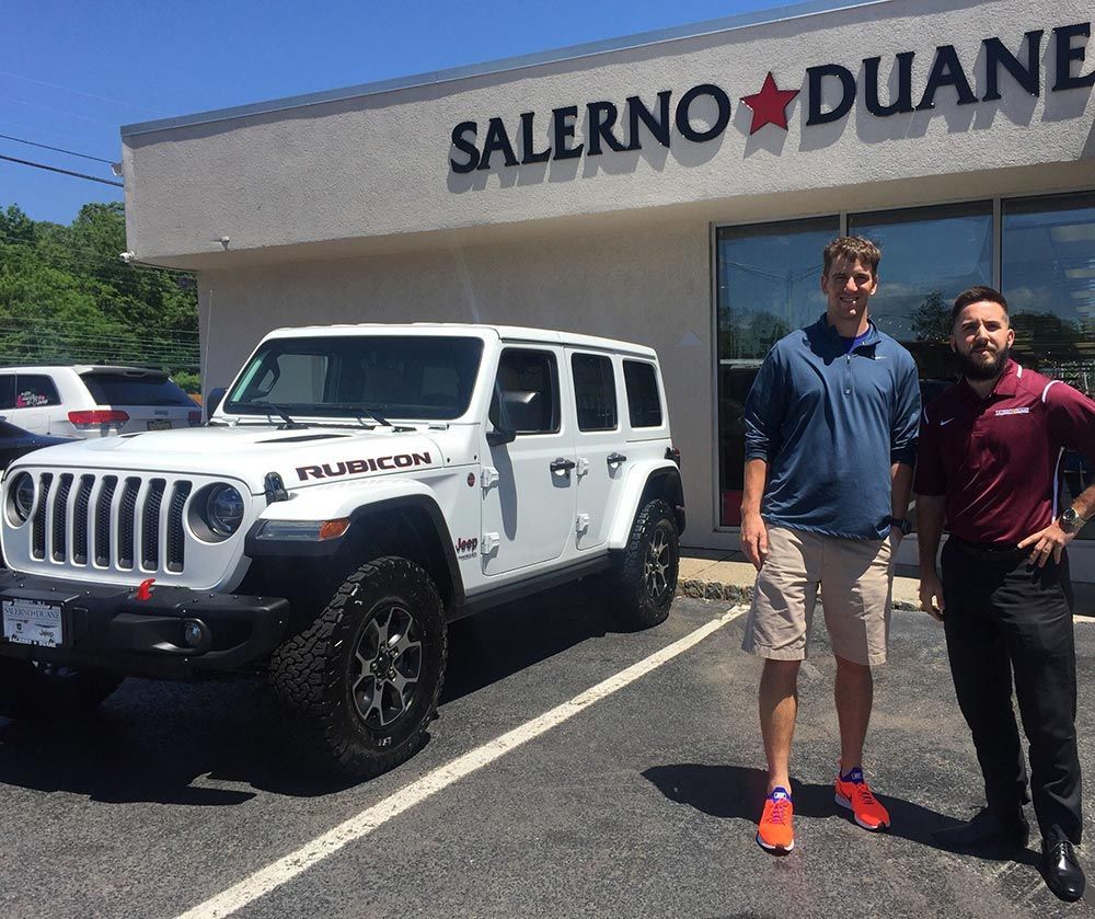 Two men are standing in front of a white jeep in a parking lot.