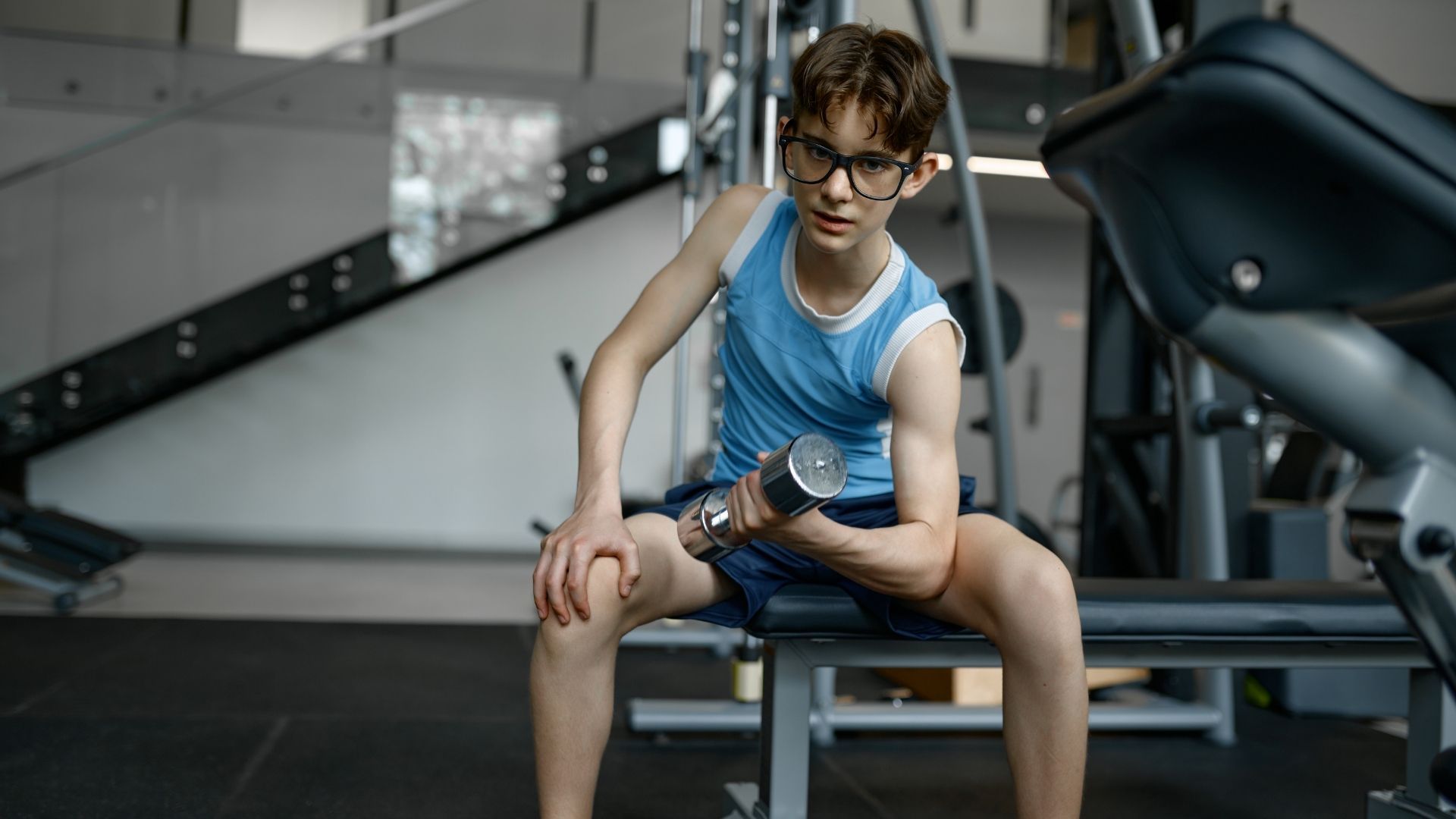 Child sitting on a gym bench, resting between dumbbell curls in a fitness center.