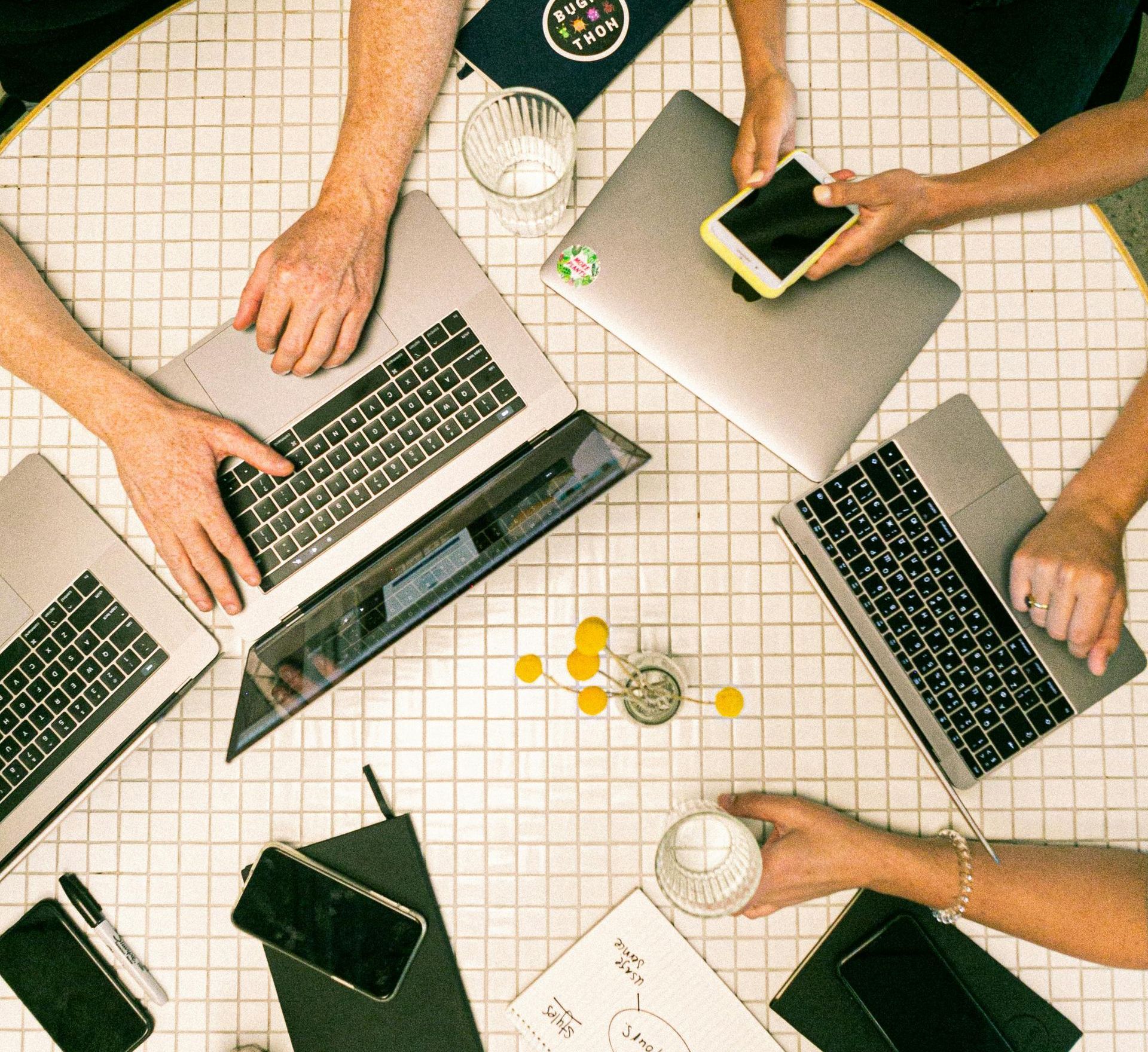 A group of people are sitting around a table with laptops and cell phones.