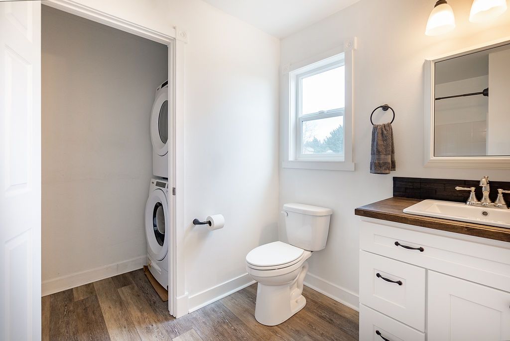 Bathroom with white vanity, toilet, and stacked washer/dryer; wood-look floor; window.