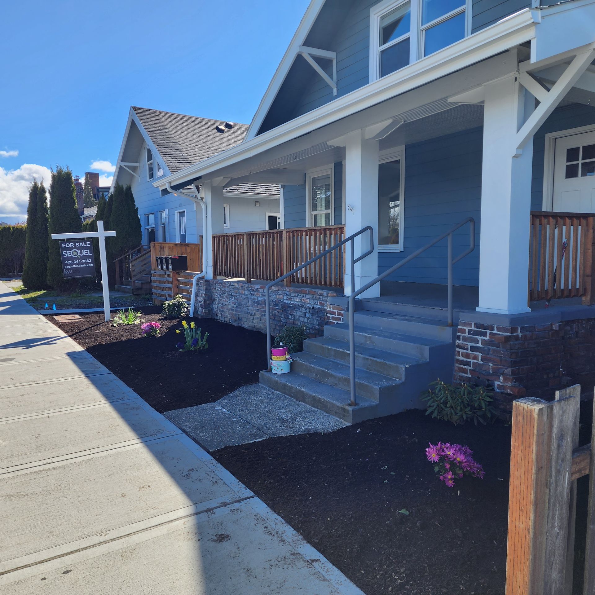 Blue house with porch, stairs, and a garden. Next to a sidewalk. Sunny day.