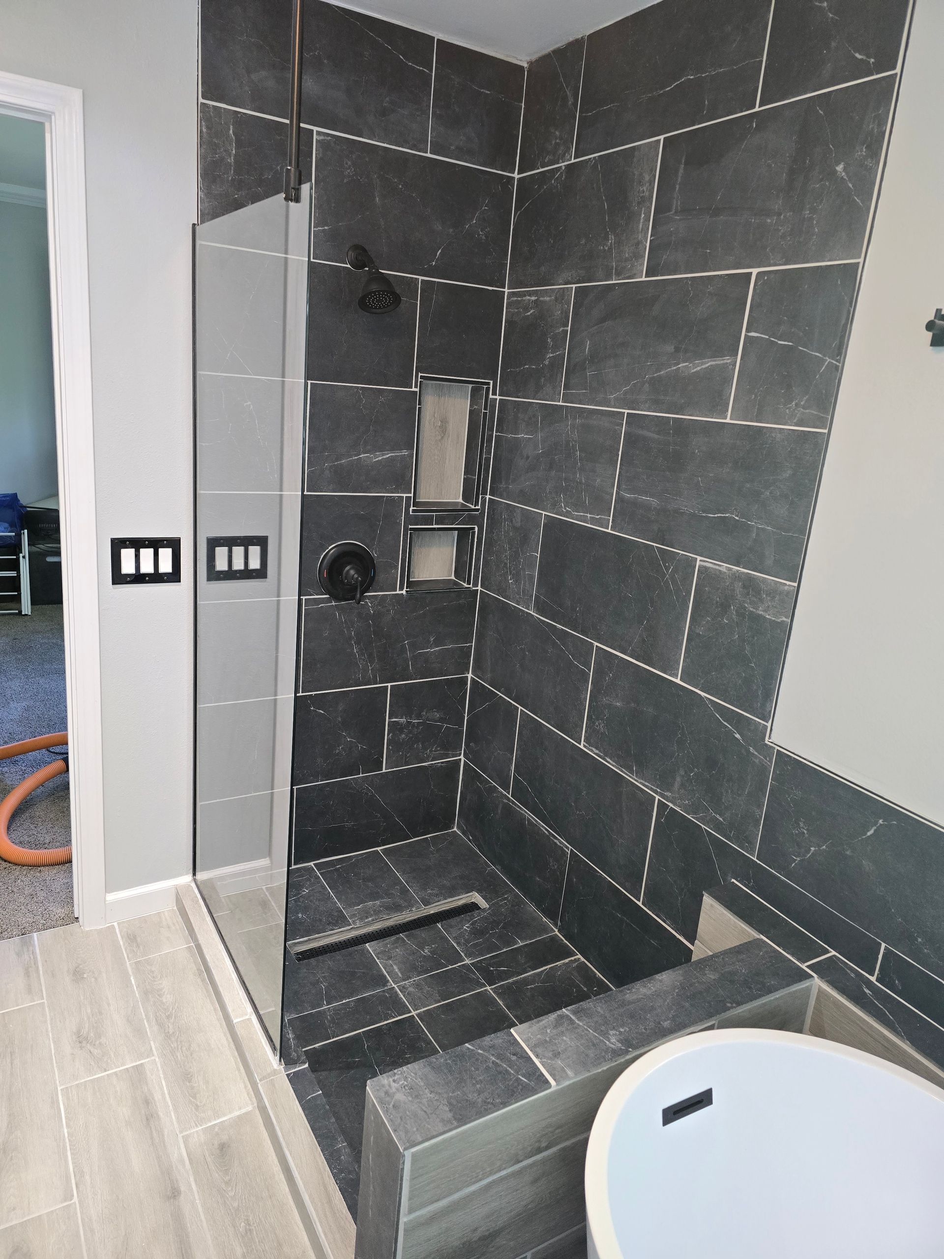A walk-in shower with dark gray tile, a glass door, and built-in shelves.