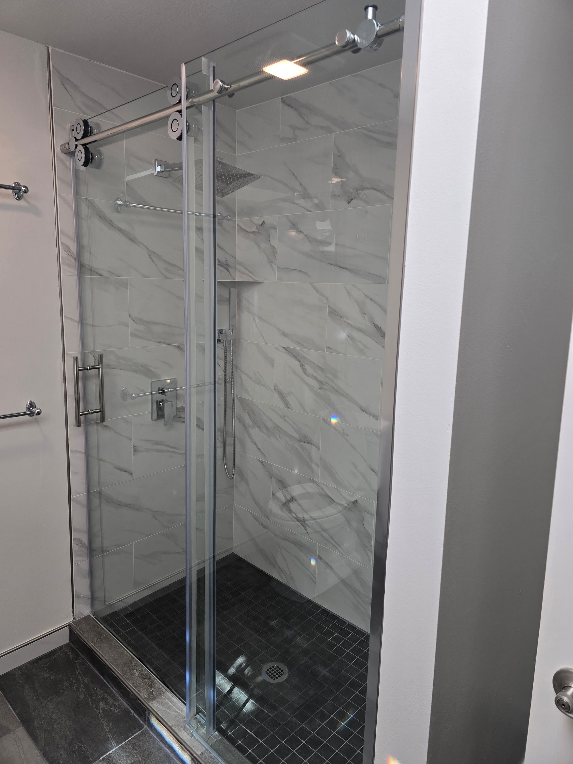 A glass-walled shower with sliding door and marble-like tile. Black shower floor and silver hardware.