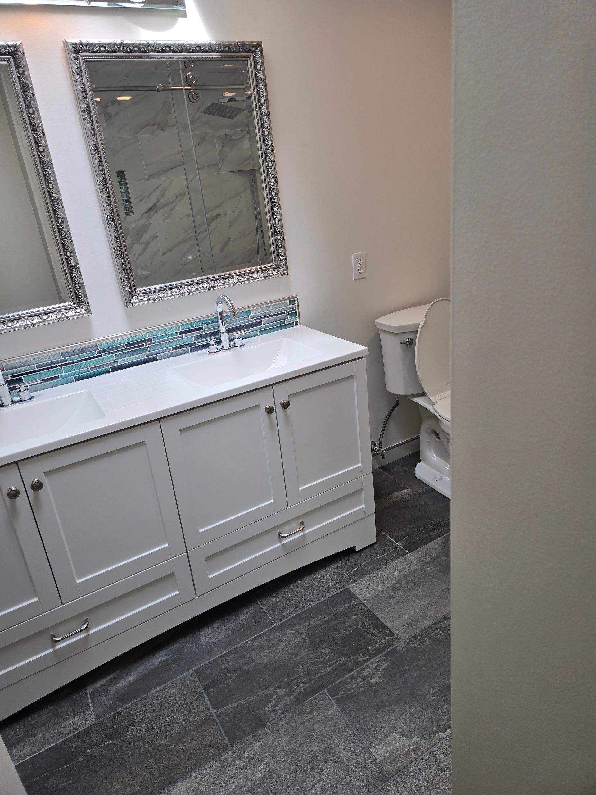 White bathroom vanity with blue tile accents, mirrors, and a toilet. Dark grey flooring.