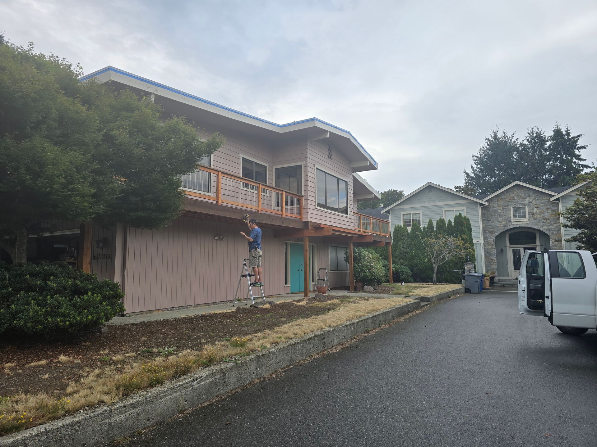 Person painting exterior of a two-story house. Cloudy day. House is pink with blue trim, and a tan wooden deck.