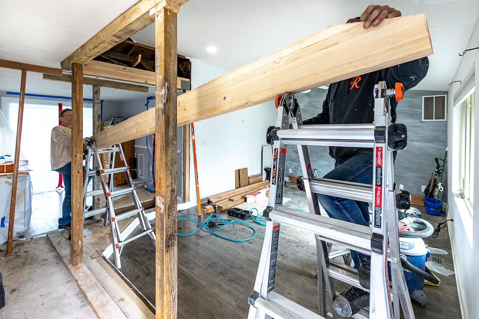 A man is standing on a ladder in a room holding a piece of wood.