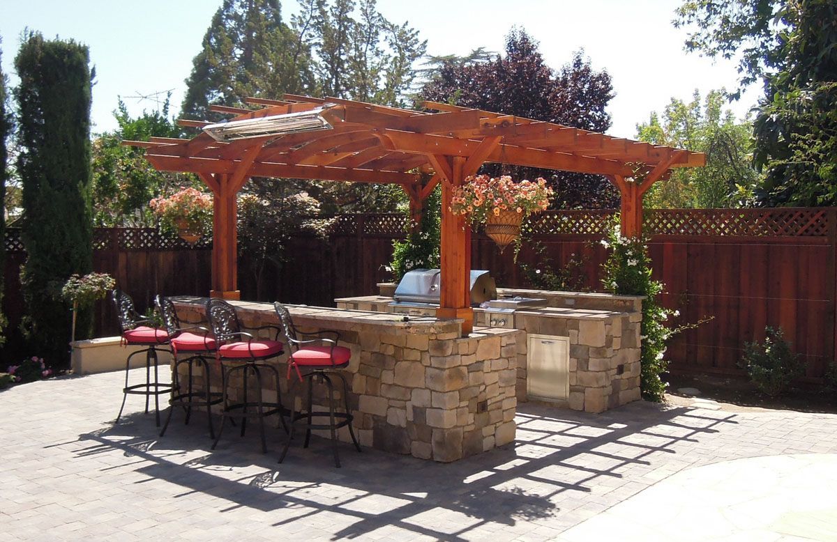 Outdoor kitchen with stone counter, red barstools, and wooden pergola.