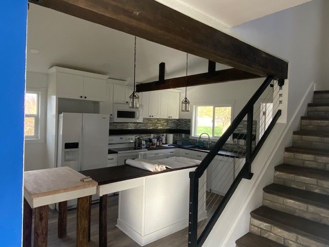 Kitchen with white cabinets, dark island, wooden beams, and staircase with black railing.
