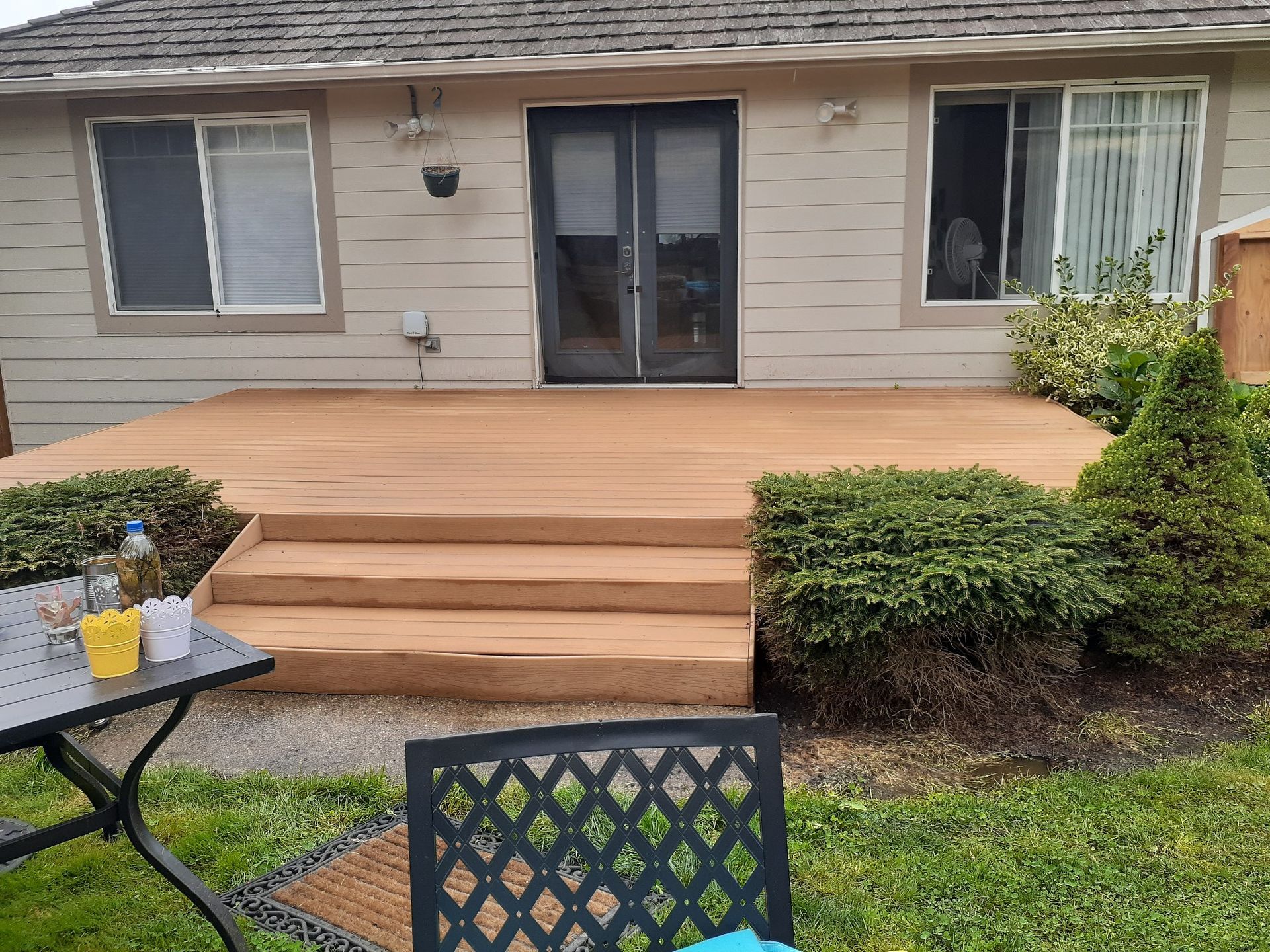 Backyard deck with steps, door, and windows on a beige house. Brown deck, green bushes, and grass in the yard.