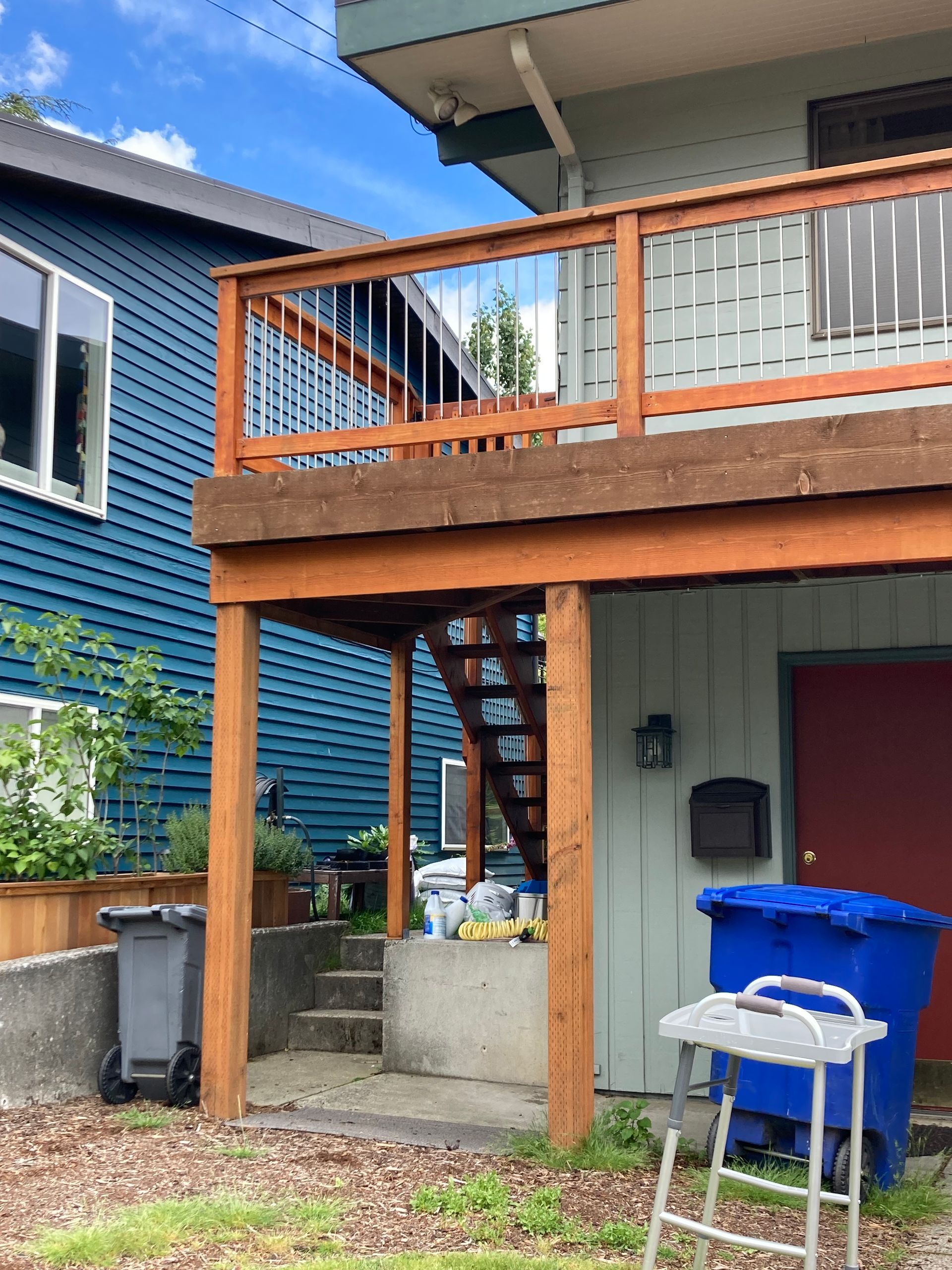 A two-story house with a wooden deck and stairs. Blue house beside it. Trash cans and a step ladder.