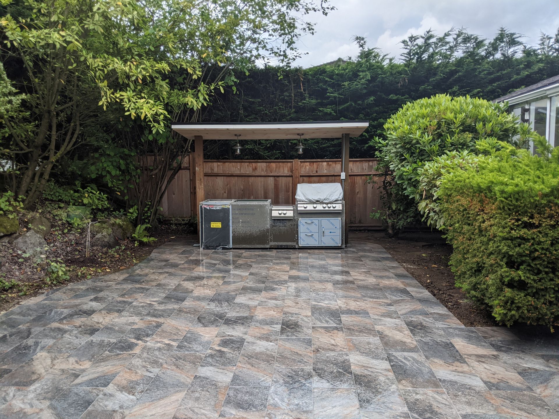 Outdoor grill area with a paved patio, wooden canopy, and surrounding greenery.