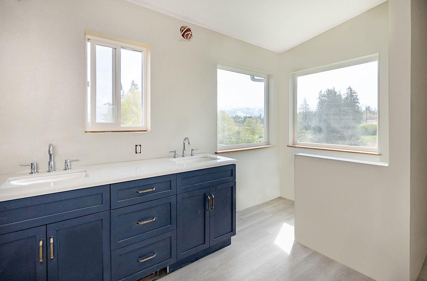 A bathroom with two sinks , two windows and blue cabinets.