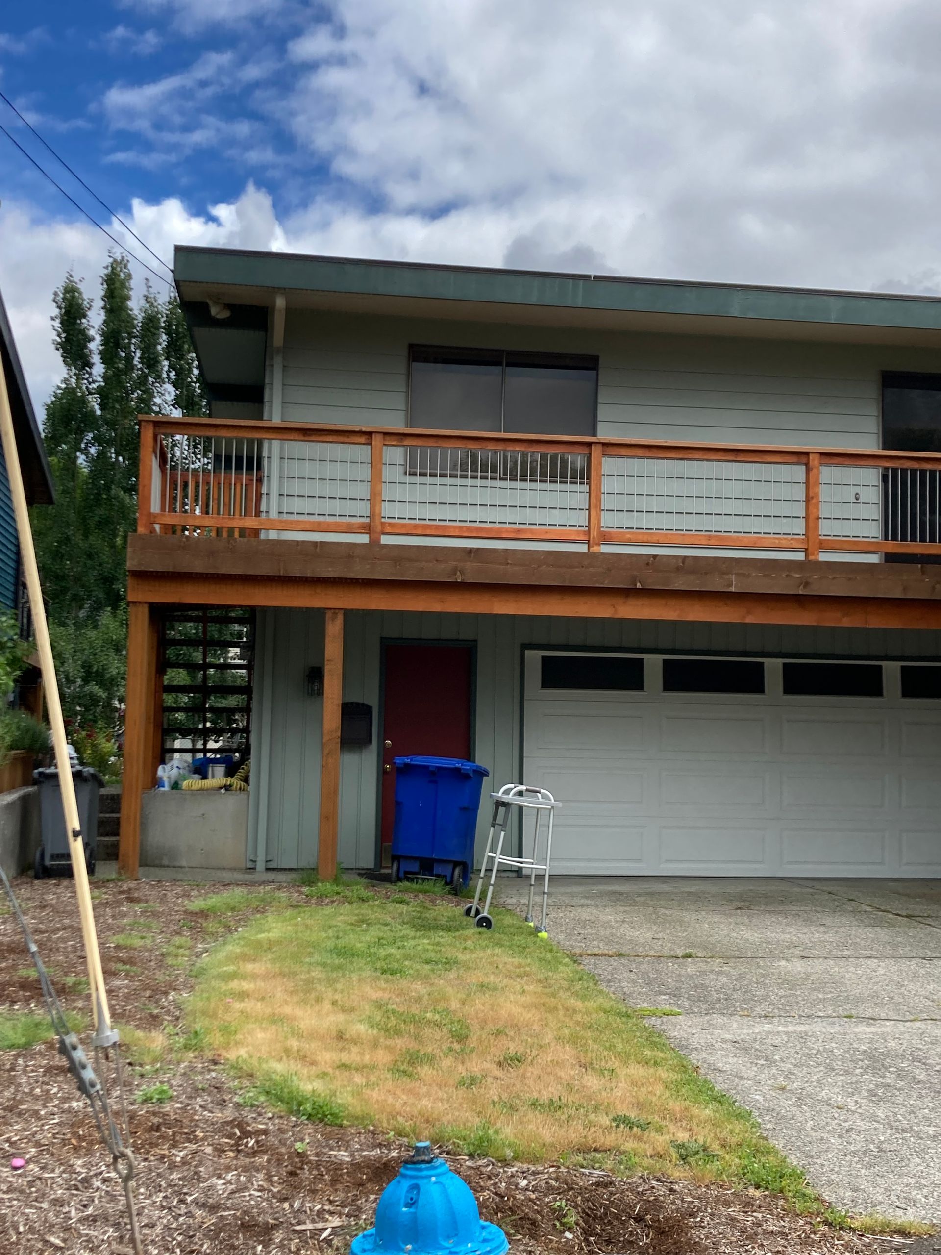 Two-story house with a deck and garage, blue trash bin, red door, blue fire hydrant, and cloudy sky.
