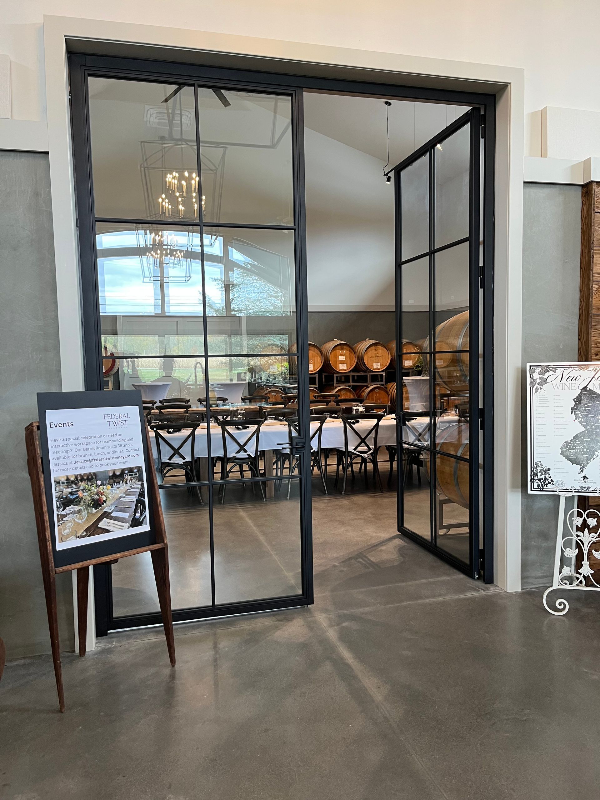 Glass doors open to a dining area with tables and barrels; an easel sign with a menu stands in front.