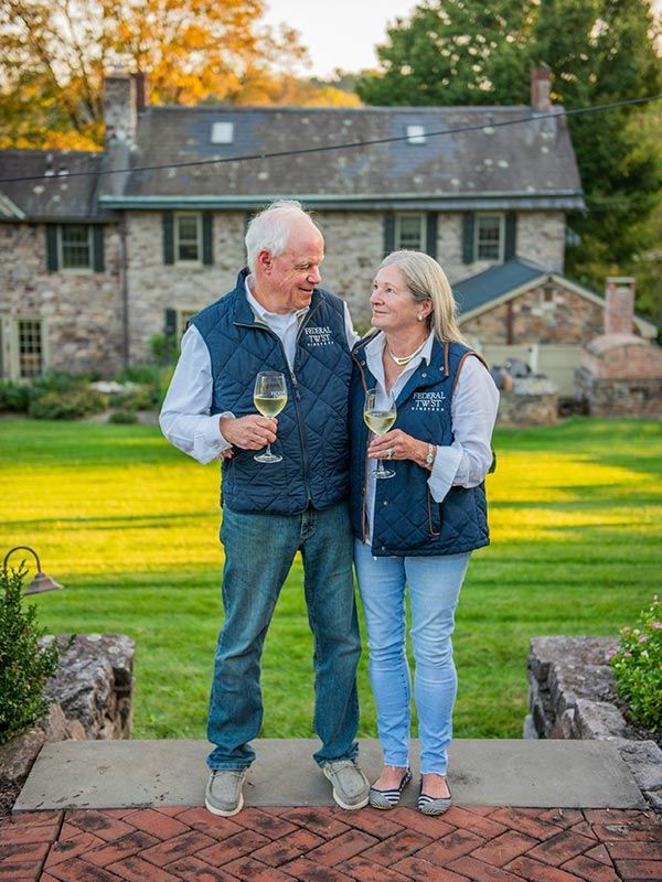 Couple in matching vests holding wine glasses, smiling at each other in front of a stone house.