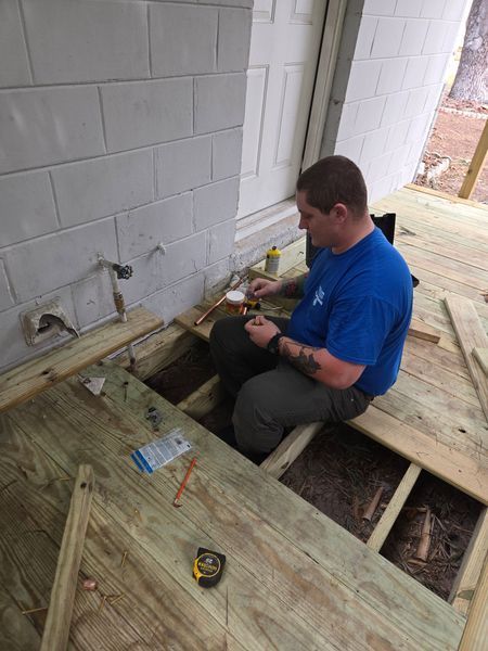 A person in blue shirt works on wooden deck, surrounded by tools and boards. Near a concrete wall.
