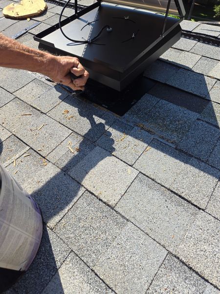 Person installing a black roof vent on gray asphalt shingles.