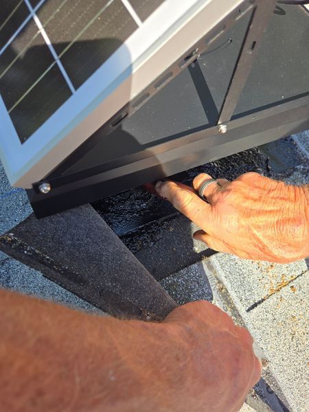Hands installing solar panel bracket onto a dark roof, pointing to screw.