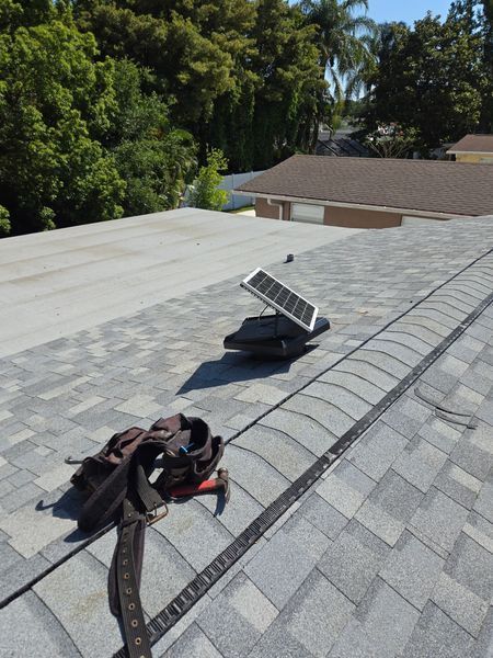 A tool belt lies on a residential asphalt shingle roof near a small solar-powered attic vent fan.