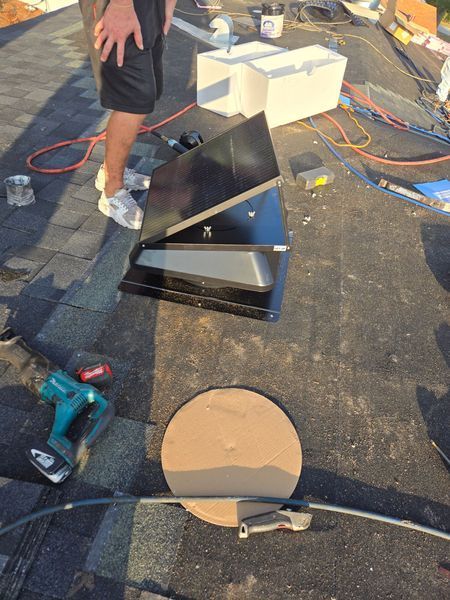 Man on a roof installing a black vent. A teal drill and circular cardboard are nearby.