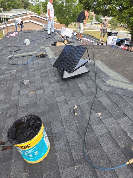Roofers working on a residential roof, with shingles, tools, and a bucket of roofing tar.