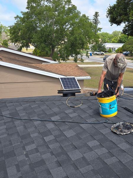Person on a roof near a solar panel and a bucket; shingles visible. A green tree and houses are in the background.