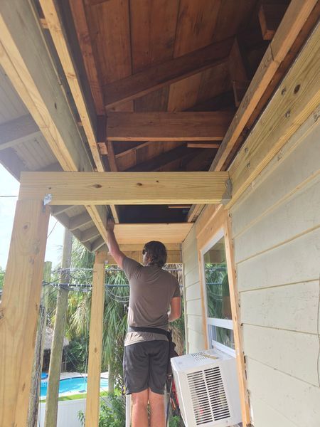 Man on ladder painting wooden porch under construction; sunlight.