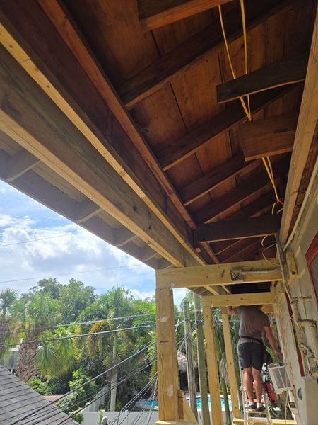 Man on ladder working on a wooden porch roof; a partially constructed structure with exposed beams and wiring.