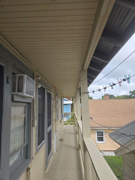 A narrow, covered outdoor walkway with windows, a railing, and a view of other houses.