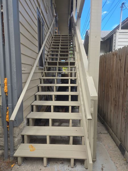 Exterior wooden staircase leading upwards, with handrails, against a light-colored building wall.