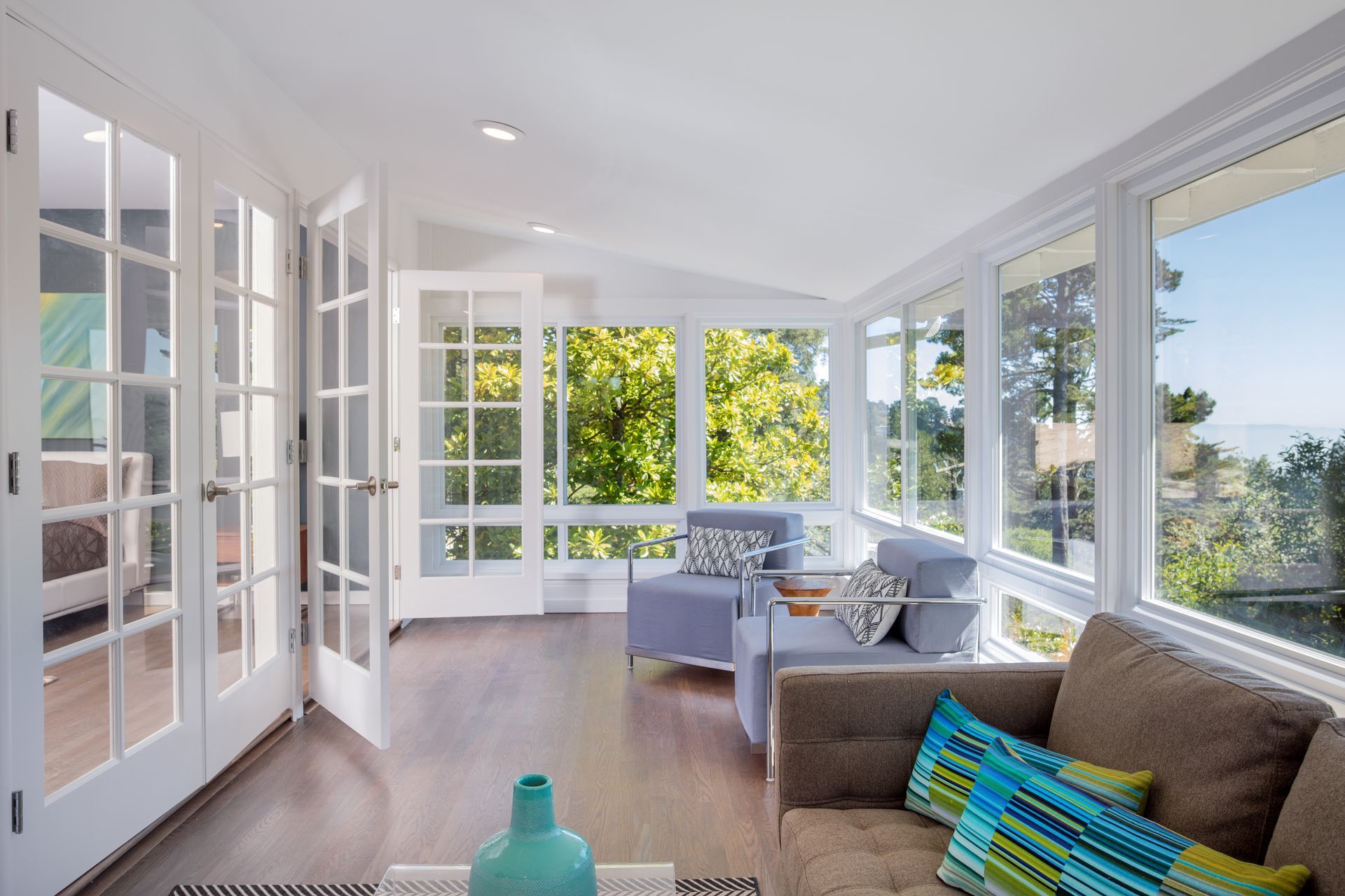 Sunroom with white-framed windows and doors, blue armchairs, and a brown sofa. Bright, sunny view of trees.
