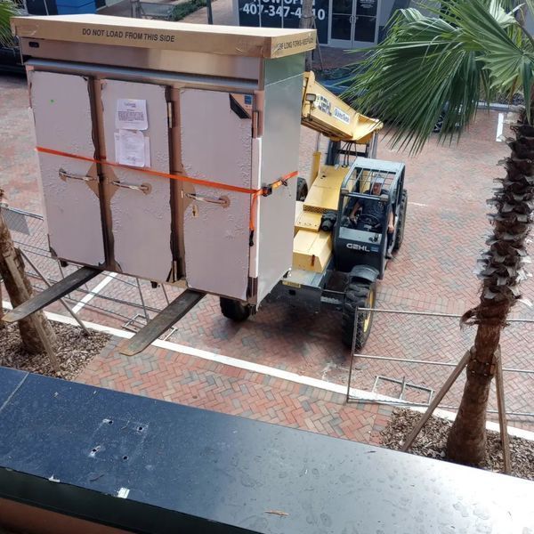 Forklift transporting a large, boxed object, possibly equipment, in an outdoor setting.
