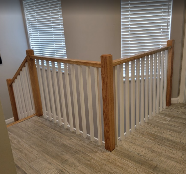 Staircase with white spindles and wooden handrails, next to windows with blinds; gray walls and flooring.