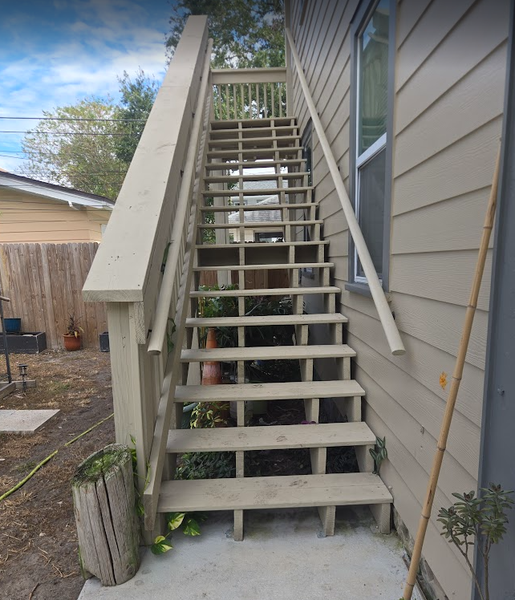 Wooden outdoor staircase leads up the side of a building, with tan siding and a simple railing.