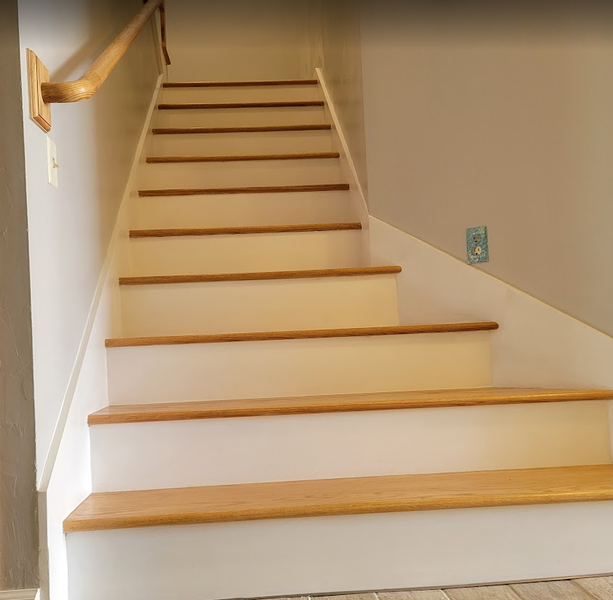 Staircase with light wood treads, white risers, and a wooden handrail against a gray wall.