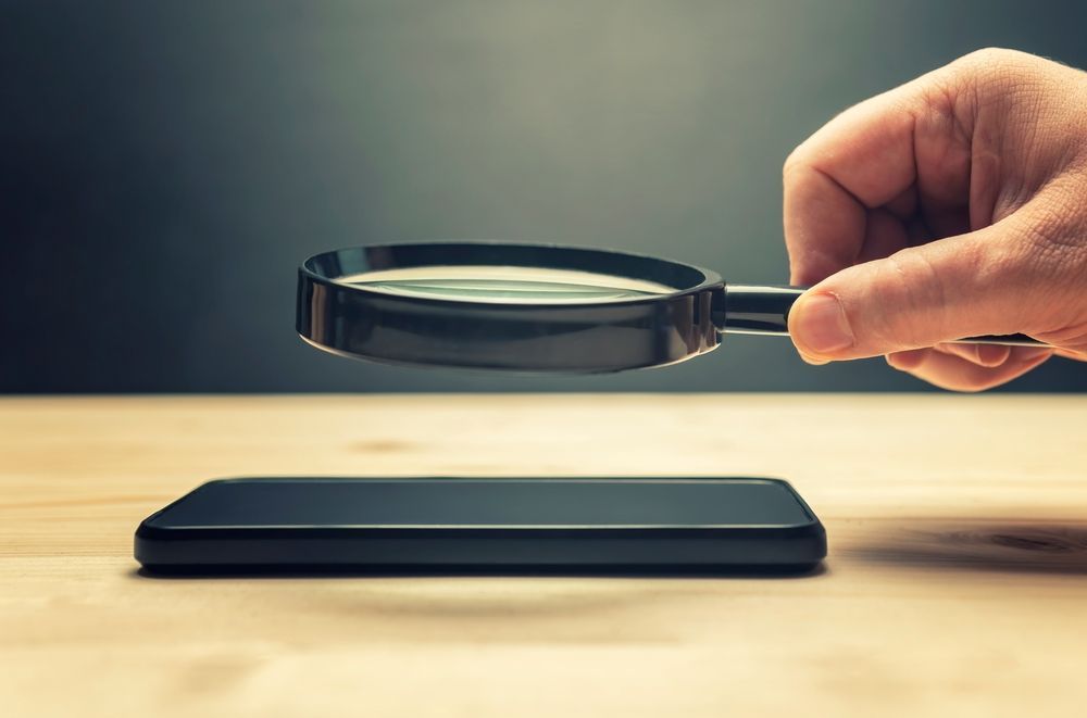 Hand holding a magnifying glass over a black smartphone on a wooden surface.