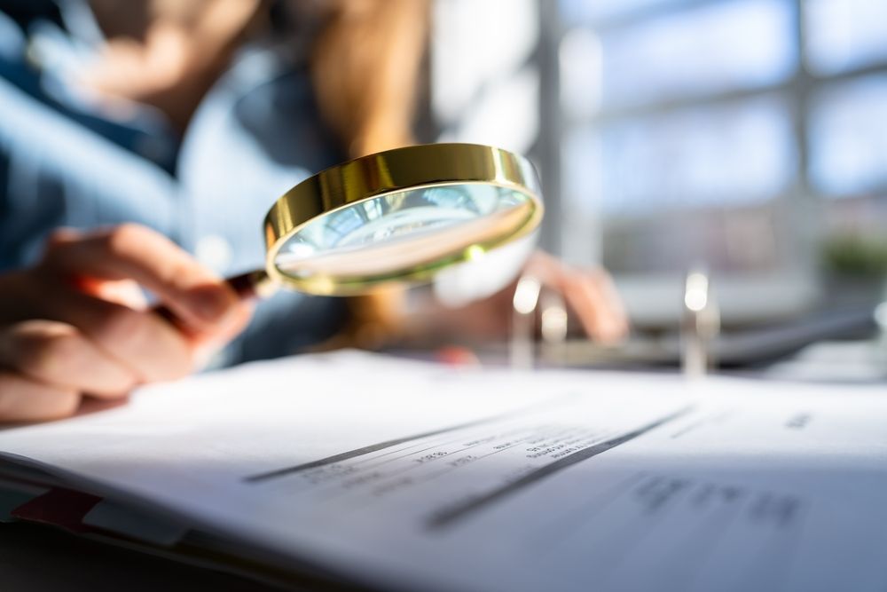 Person examining document with magnifying glass; sunlight in the background.