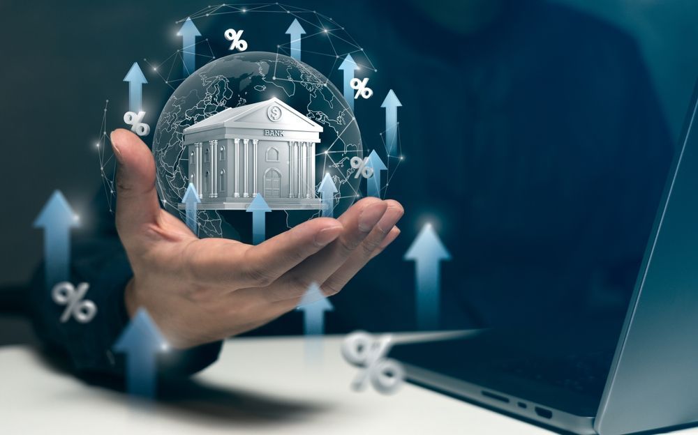 Hand holding a bank, surrounded by upward arrows and percentage signs, next to a laptop.