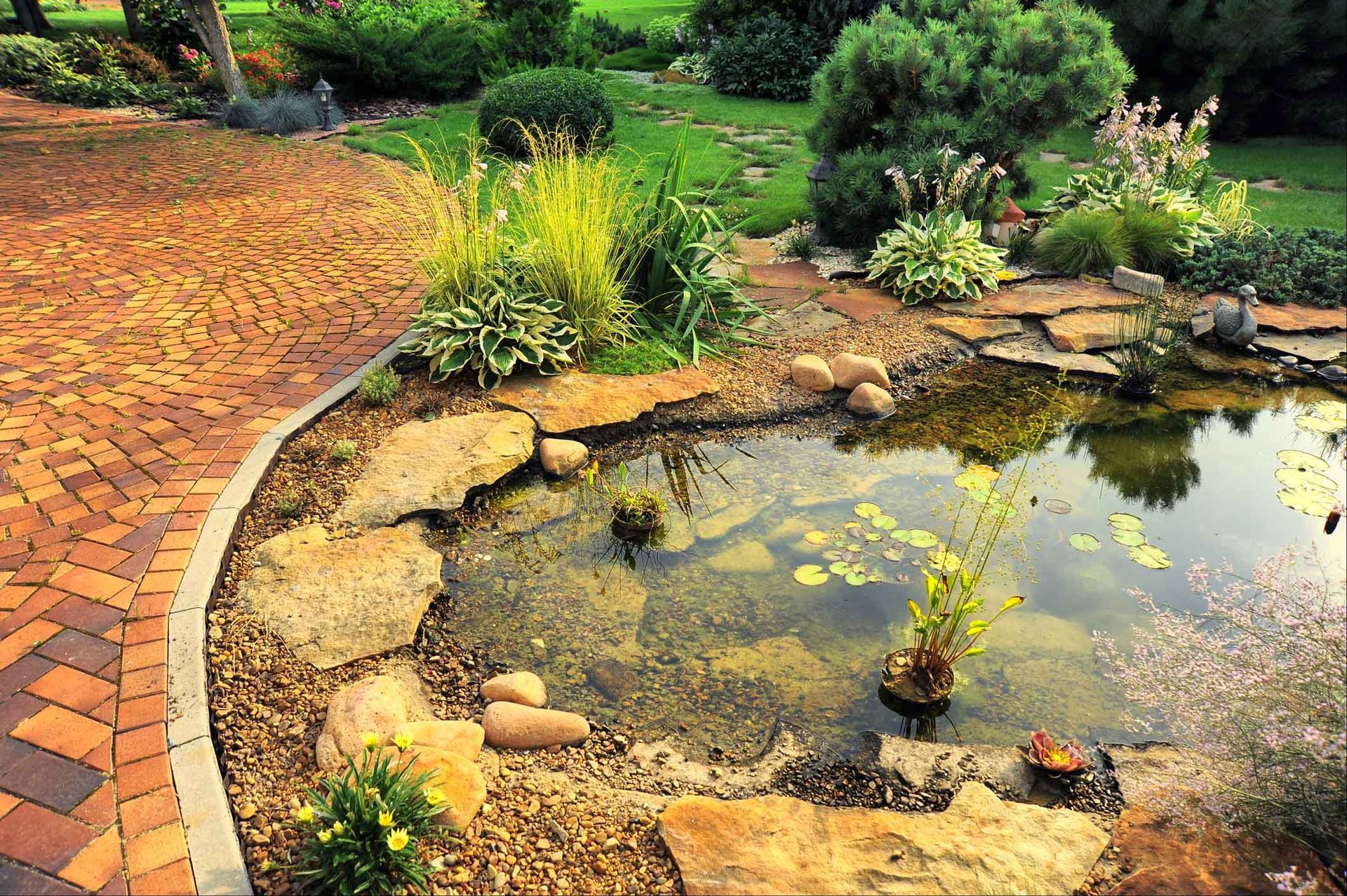 A stone-edged pond surrounded by lush greenery and flowers next to a curved, multi-colored brick patio. A stone-edged pond surrounded by lush greenery and flowers next to a curved, multi-colored brick patio.